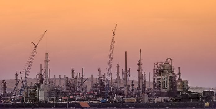 Building Oil or Natural Gas Refinery with Construction Cranes against an orange sunset background.