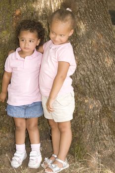 Two Young Ethnic Sisters with Questioning and Mischievous looks on their faces. They have their arms around each other as hold each other while standing against the trunk of a large tree.