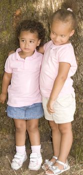 Two Young Ethnic Sisters with Questioning and Mischievous looks on their faces. They have their arms around each other as hold each other while standing against the trunk of a large tree.