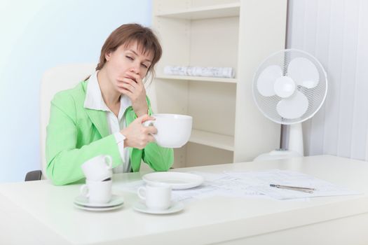 The tired woman sits at a table with the big cup of coffee and yawns