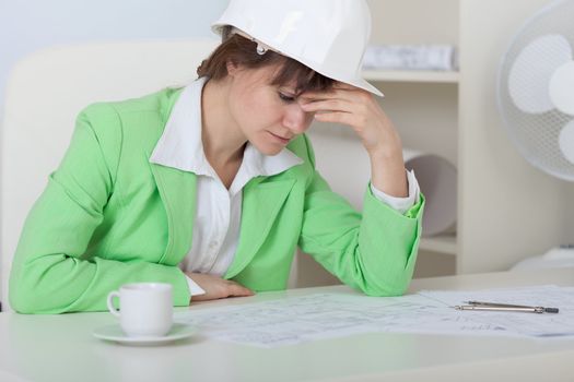 Tired woman - the engineer in a helmet sits at a table at office