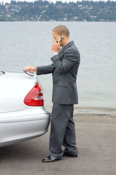 Ethnic Business Man Making a Sale on Cell Phone holding a calculator while standing by his luxury car at the lake. 