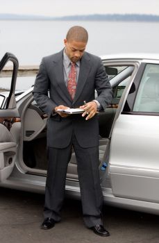 Ethnic Business Man with Clipboard and Pen next to Luxury Car working at the lake.