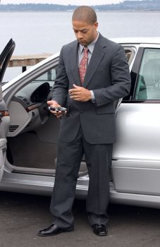 Left handed ethnic business man using a PDA next to his luxury car parked at the lake. He is wearing a suit and tie.