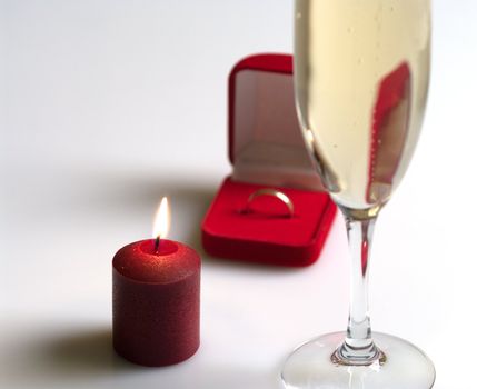 Small box with a gold ring, tall wine glass of champagne and conflagrant candle. Focus on a candle