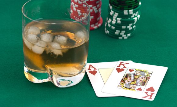 Pile of counters, playing cards and glass of whisky with ice on green cloth.