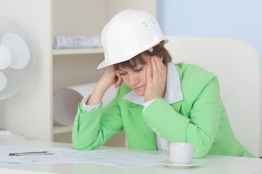 Woman the engineer in a protective helmet sits at a table on a workplace