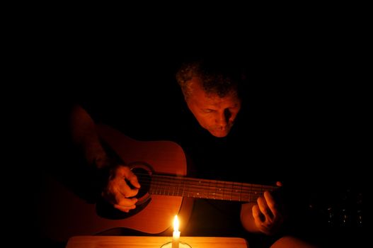 A man playing his guitar at nightime with just the light of a single candle.