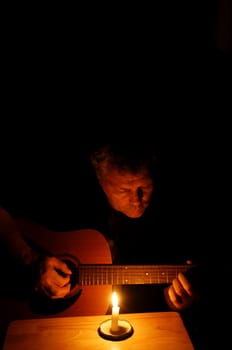 A middle-aged man playing his guitar at night in the glow of a lit candle.