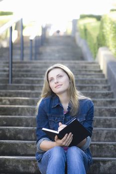 Pretty Blonde Teen Girl Sitting on a Stairway Outdoors Writing in Journal