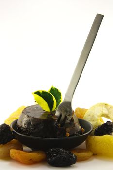 Small christmas pudding in a black dish with assorted dried fruit and a small silver fork on a reflective white background