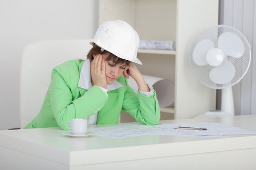 Sad woman the engineer with a helmet on a head sits at a table on a workplace