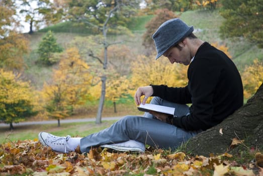 Young man reading under a tree in the park during autumn.