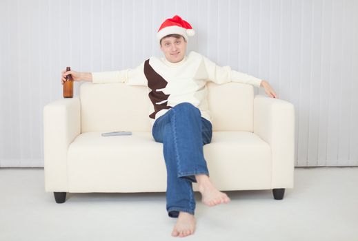 The person in a Christmas cap comfortably sits on a sofa with a beer bottle