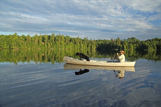 Canoeist with a black Labrador Retriever in the bow paddling on a northern Ontario lake with beautiful reflection on the water