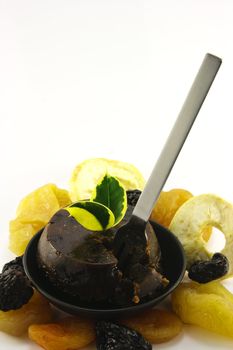 Small christmas pudding in a black dish with assorted dried fruit and a small silver fork on a reflective white background