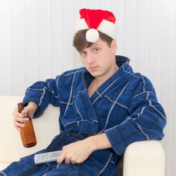 The man in a Christmas cap sits on a sofa in a dressing gown with remote control and beer