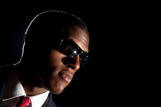 Low key portrait of an African American business man dressed in a suit and sunglasses standing in front of a dark black background.