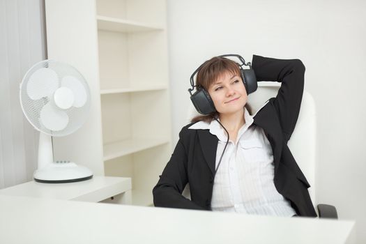 The young beautiful woman sits at office with ear-phones on a head and listens to music