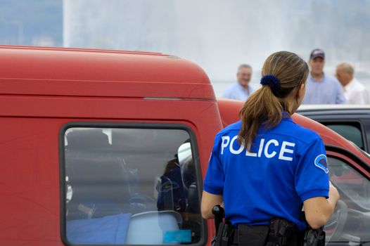 A female gendarme questions a motorist in an old, beat-up van as onlookers watch from a distance. Space for text in the sky above the van.