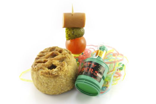 Small pork pie with party popper and cocktail stick containing hot dog sauage, gherkin and tomato with streamers on a reflective white background