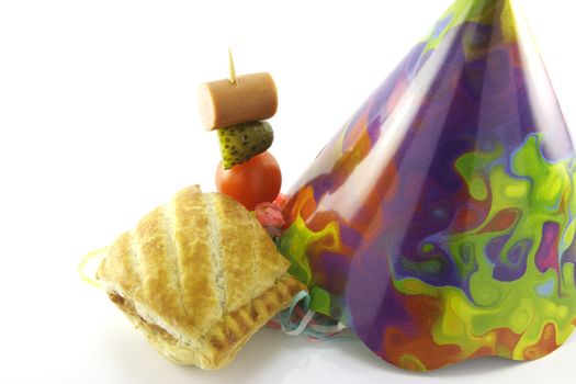 Small sausage roll with cone party hat and cocktail stick containing hot dog sausage, gherkin and tomato on a reflective white background