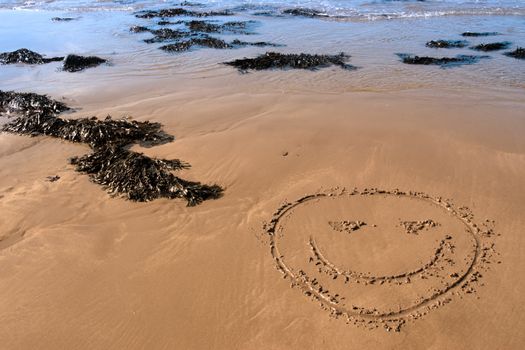 a smiley face icon inscribed on the beach with waves in the background on a hot sunny day
