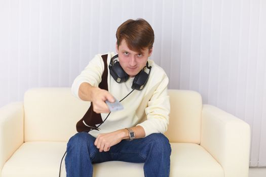The man sits on a sofa with earphones and remote control