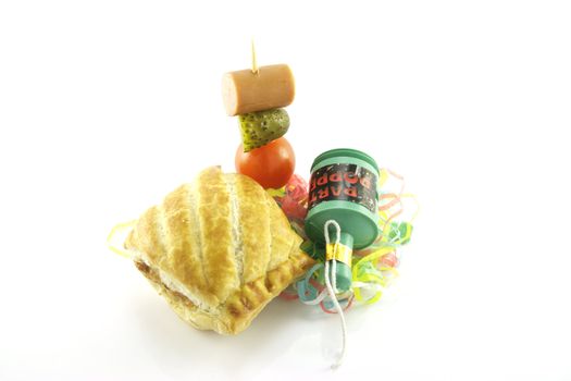 Small sausage roll with party popper and cocktail stick containing hot dog sausage, gherkin and tomato with streamers on a reflective white background