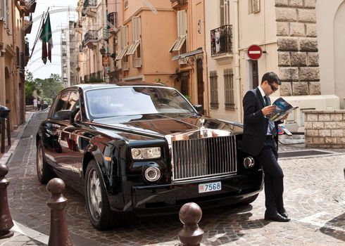 Monaco, chauffeur waits by Rolls Royce reading newspaper in the street of the tax haven city.