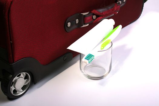 The tooth-paste and tooth-brush lie on a pure glass. On a back background a suitcase of red colour.