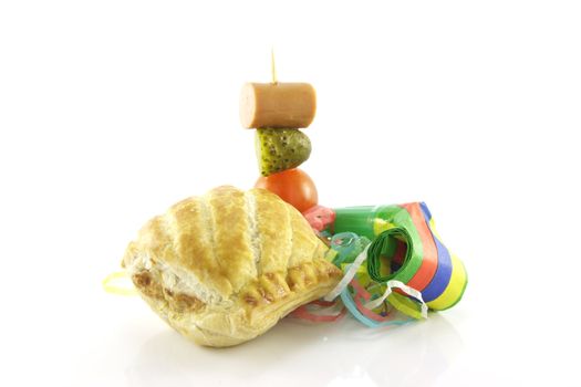 Small sausage roll with party blower and cocktail stick containing hot dog sauage, gherkin and tomato on a reflective white background