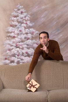 A secret santa is leaving behind his gift on a sofa and telling the viewer to be quiet
