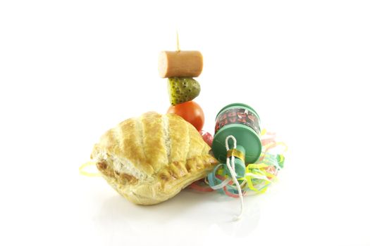 Small sausage roll with party popper and cocktail stick containing hot dog sausage, gherkin and tomato with streamers on a reflective white background