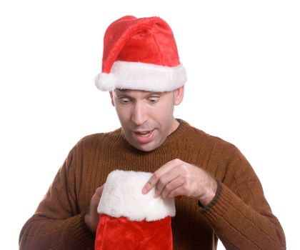 A young man wearing a Santa hat is looking inside his Christmas stocking and looks surpised, isolated against a white background