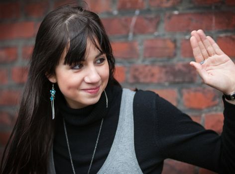 Smiling brunette girl greets somebody with raised hand; brick wall in background