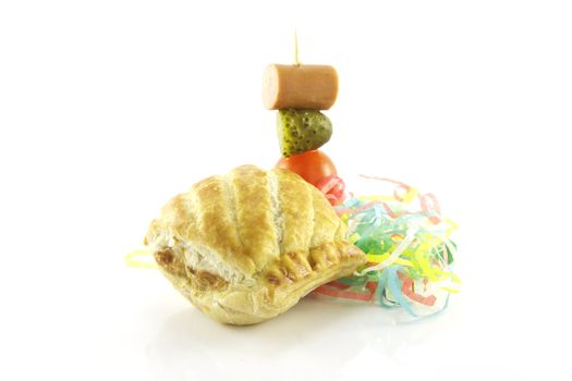 Small sausage roll with cocktail stick of hot dog sausage, gherkin and tomato with streamers on a reflective white background