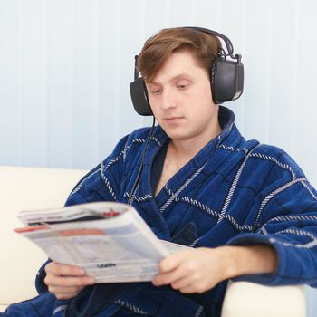 The man in the big ear-phones sits on a sofa reads the newspaper