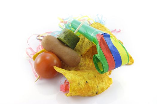 Baked spicy nachos with cocktail stick containing tomato, hot dog and gherkin with blower and party streamers on a reflective white background