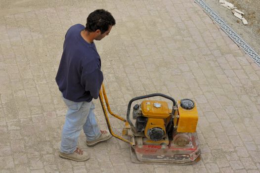 A builderusing a machine to pack down bricks in a newly-paved courtyard. A little of the rest of the building site can be seen at the top right.