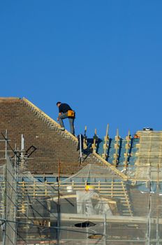 A workman, high on a roof, above the scaffolding, with his orange tool bag attached to his belt, making repairs.