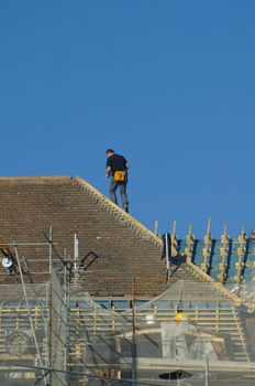 A workman, high on a roof, with an orange tool bag attached to his belt. Roof under repair.