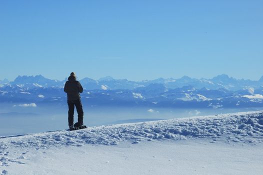 A skier, high on a ridge in the Swiss Jura mountains in winter, takes a break from skiing and gazes across to the French Alps. The jagged row of peaks to the left of her head are the Dents du Midi. In the valley below a line of mist can be seen, caused by an atmospheric inversion layer.