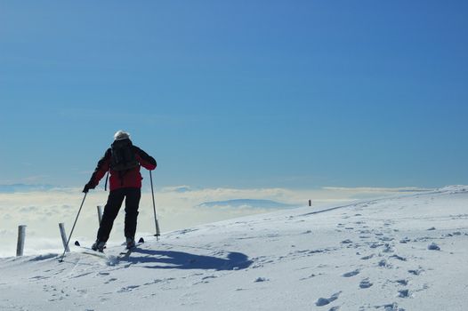 A skier sets off across a gentle snow-covered slope, high on a mountain in the Swiss Jura range. Below him the valley is filled with cloud and, far off in the distance, peaks of the Alps can be faintly seen.