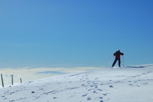 A skier skis away across a gentle snow-covered slope, high on a mountain in the Swiss Jura range. Below him the valley is filled with cloud and, far off in the distance, peaks of the Alps can be faintly seen.