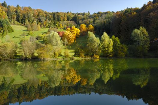 The colours of autumn in an arboretum. Taken in the early-morning light, under a clear blue sky. The flame-like colours of the trees are reflected in the stil waters of a lake. A tiny figure can be seen walking past the brightest trees.