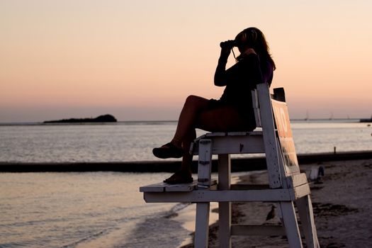 A silhouette of a woman looking with binoculars at the beach while seated in a lifeguard chair.