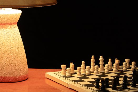 Luminous table lamp standing near chess board on dark background