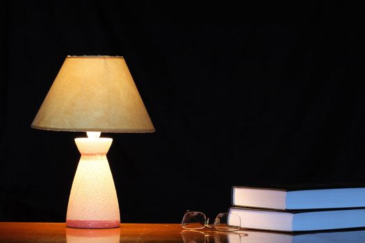 Luminous table lamp standing near spectacles and books on dark background