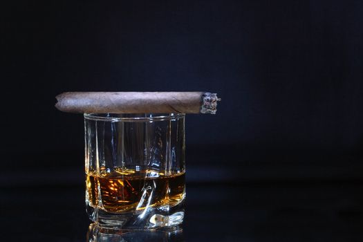 Cuban cigar lying on glass of whiskey isolated on dark background with copy space
